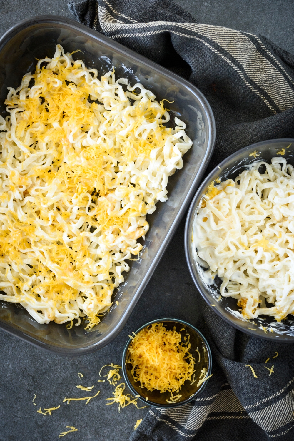 A baking dish filled with a layer of cooked noodles and shredded cheese. Next to the baking dish is a bowl of noodles in cream sauce and a bowl of shredded cheese.