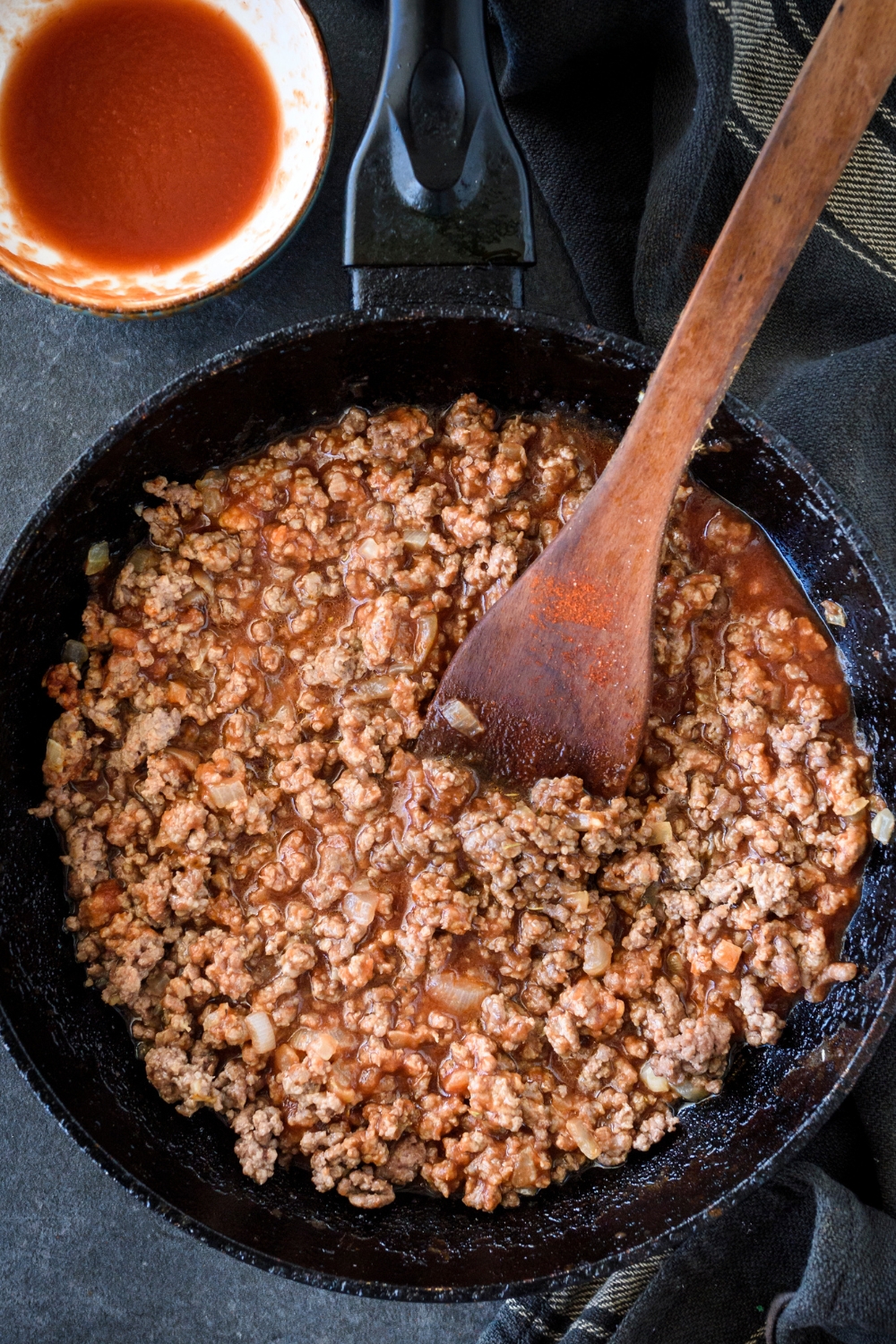 A skillet filled with cooked ground beef in tomato sauce. There is a wooden spoon in the skillet.