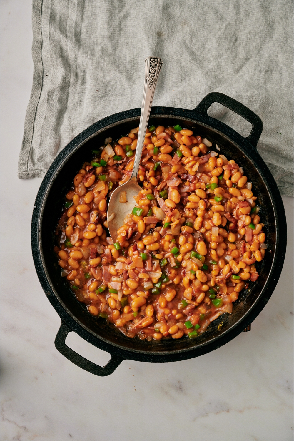 A black skillet filled with canned beans, diced bacon, diced green bell pepper, diced onion, and a spoon is in the skillet.