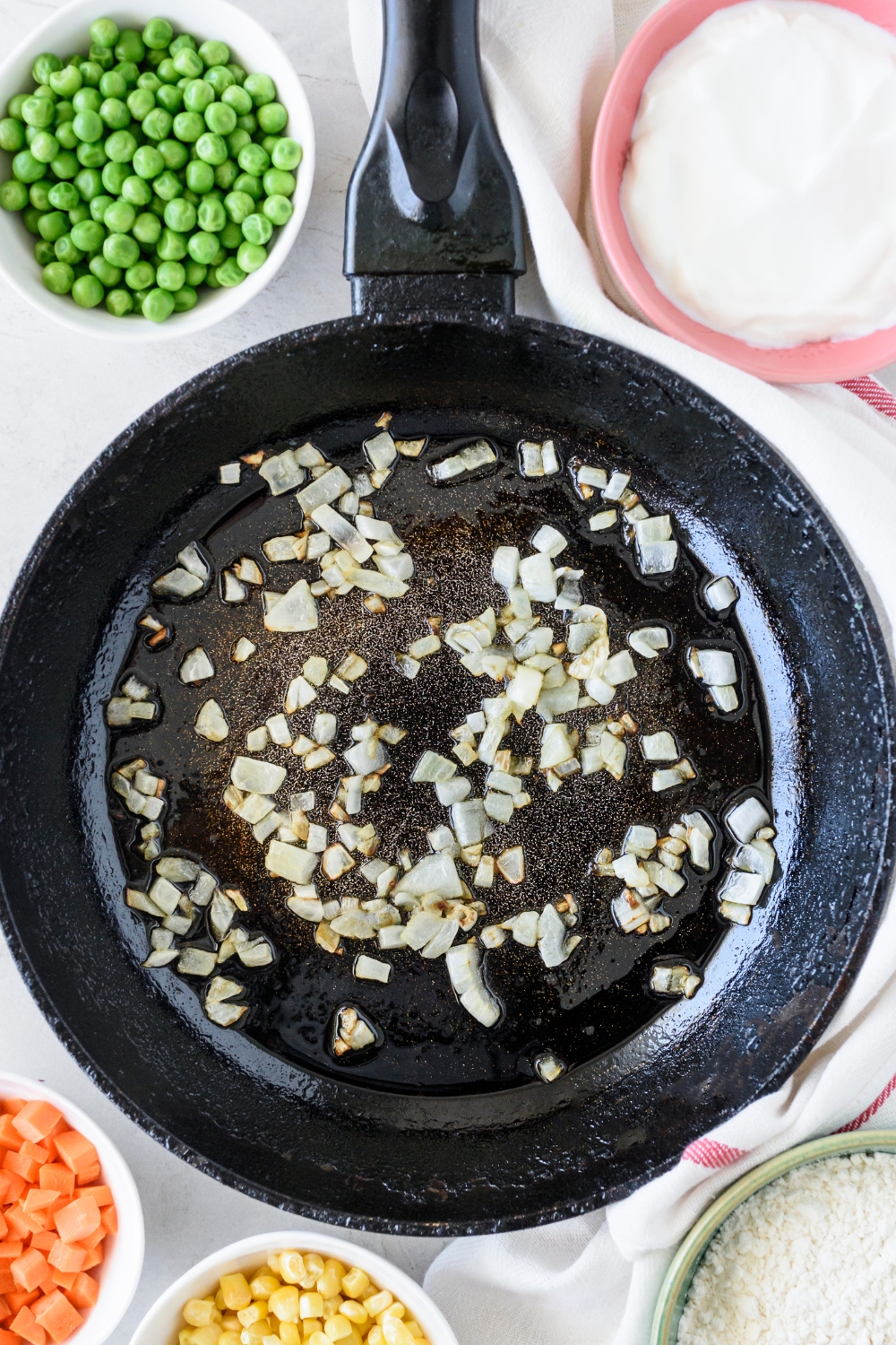 A black skillet with diced onion cooking in oil. The skillet is surrounded by an assortment of ingredients.