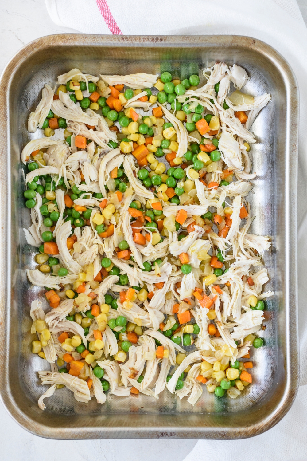 A rectangular baking dish filled with corn kernels, diced carrots, peas, and shredded chicken spread in an even layer along the bottom of the dish.