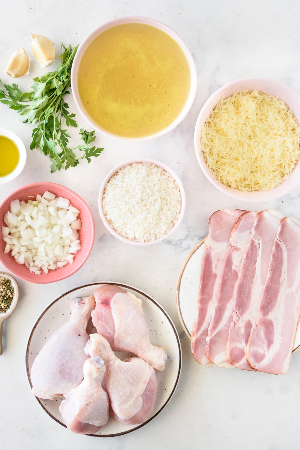 An assortment of ingredients including bowls of broth, shredded cheese, diced onion, plates of uncooked bacon and raw chicken thighs, and a bunch of parsley next to two garlic cloves.