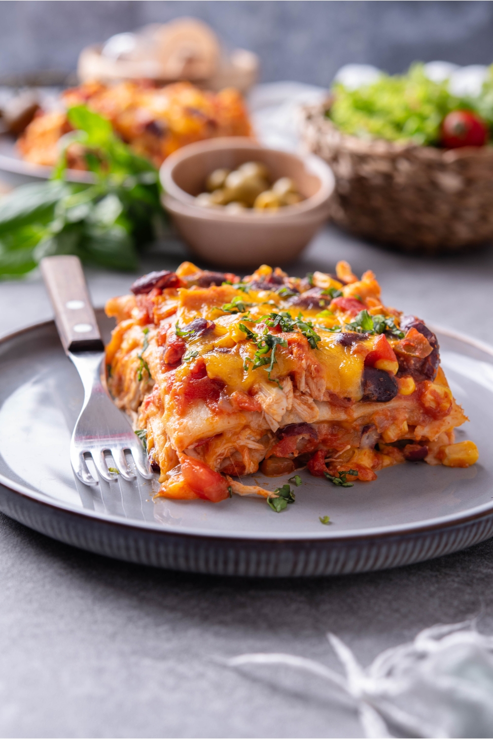 A square serving of chicken tortilla casserole layered with tomato sauce, shredded chicken, black beans, and topped with melted cheese, and fresh green herbs.