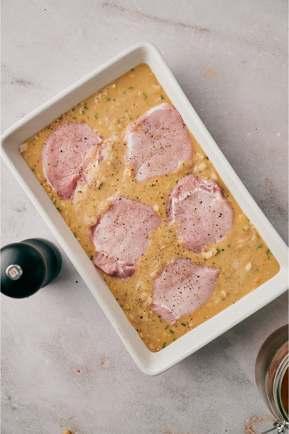 A white baking dish filled with a creamy soup mixture and five pork chops are layered on top and seasoned with salt and pepper.