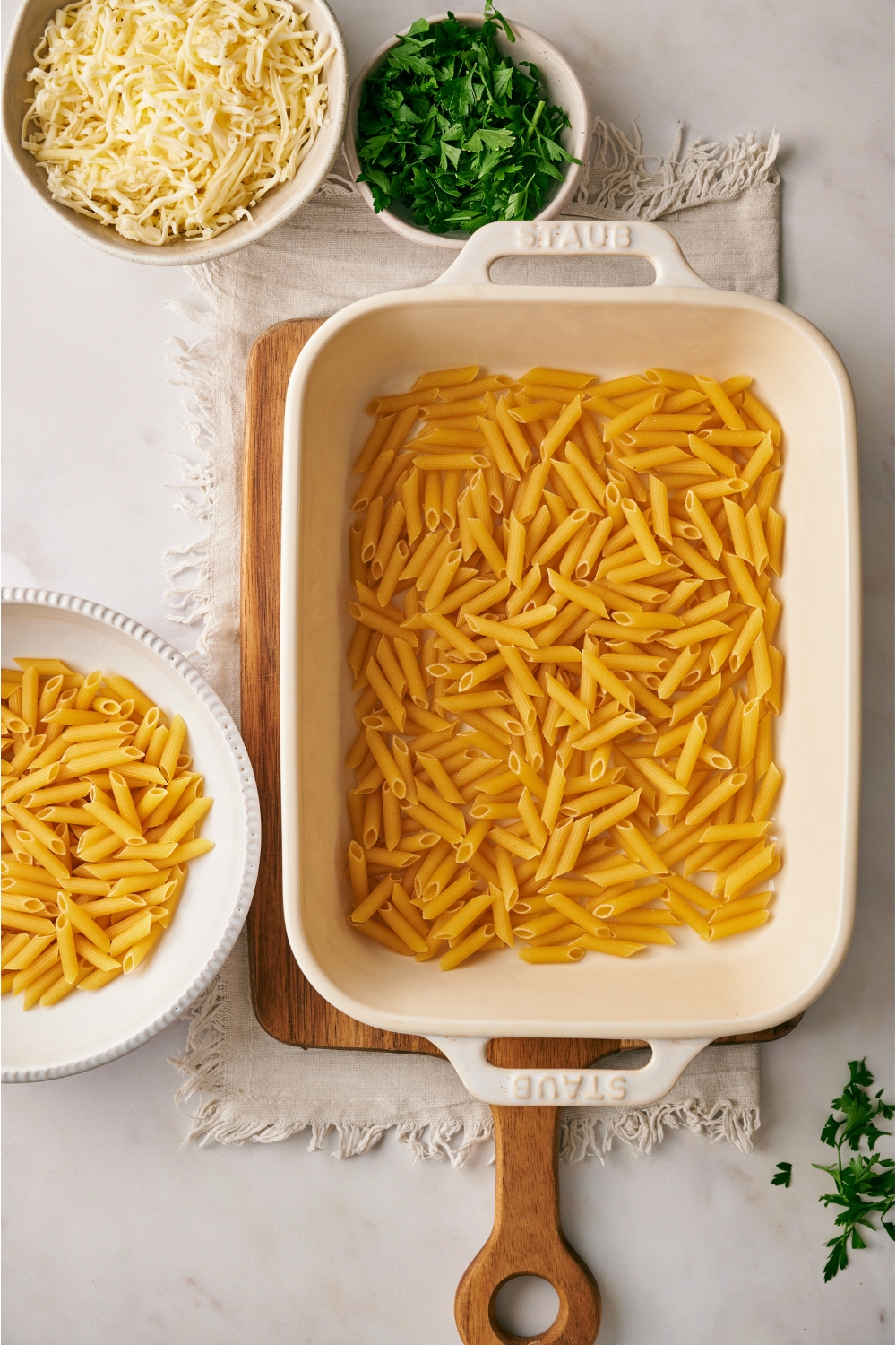 A white baking dish filled with dried penne pasta, next to bowls of dried pasta, shredded cheese, and fresh green herbs.