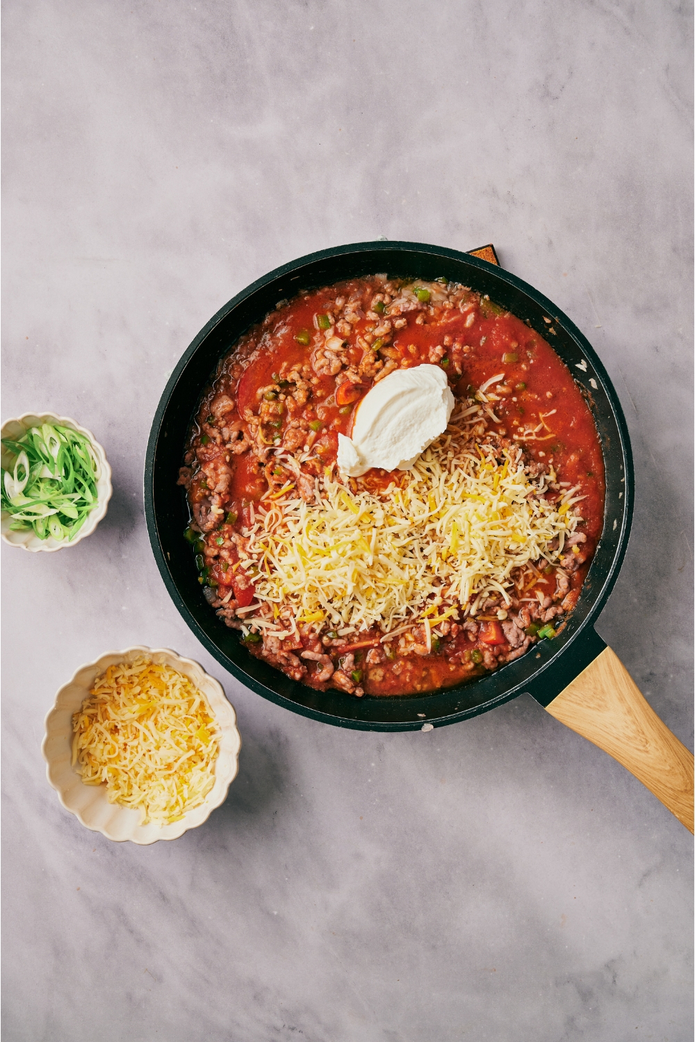 A skillet filled with ground beef, diced peppers and onion, tomato sauce, shredded cheese, and a dollop of sour cream added but not yet combined.