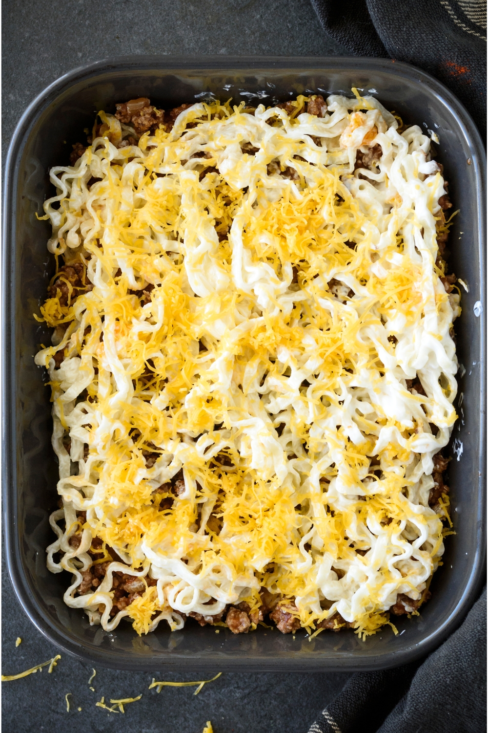 A baking dish filled with layers of cooked ground beef, cooked noodles in cream sauce, and shredded cheese on top.