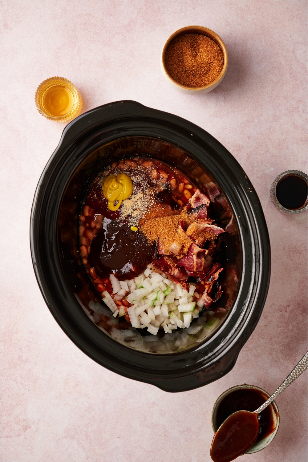 A crock pot filled with canned beans, diced onion, barbecue sauce, yellow mustard, and spices.