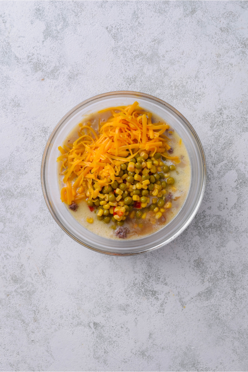 A clear bowl filled with a creamy soup mixture, mixed vegetables, and shredded cheddar cheese.