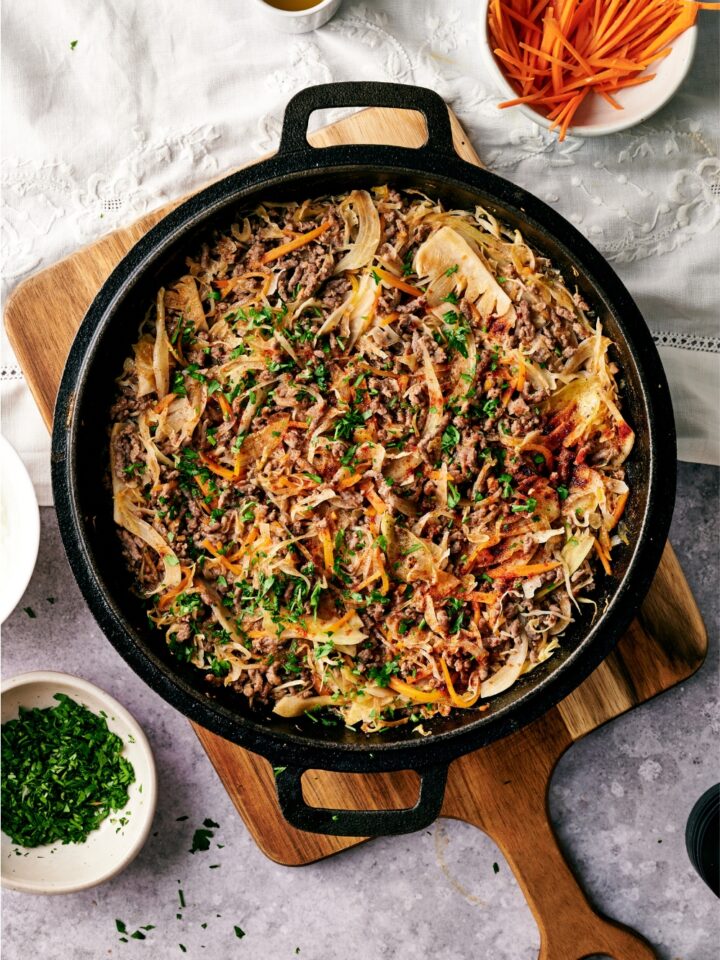 A black skillet filled with ground beef and cabbage stir fry garnished with fresh parsley. The skillet is on a wooden board.