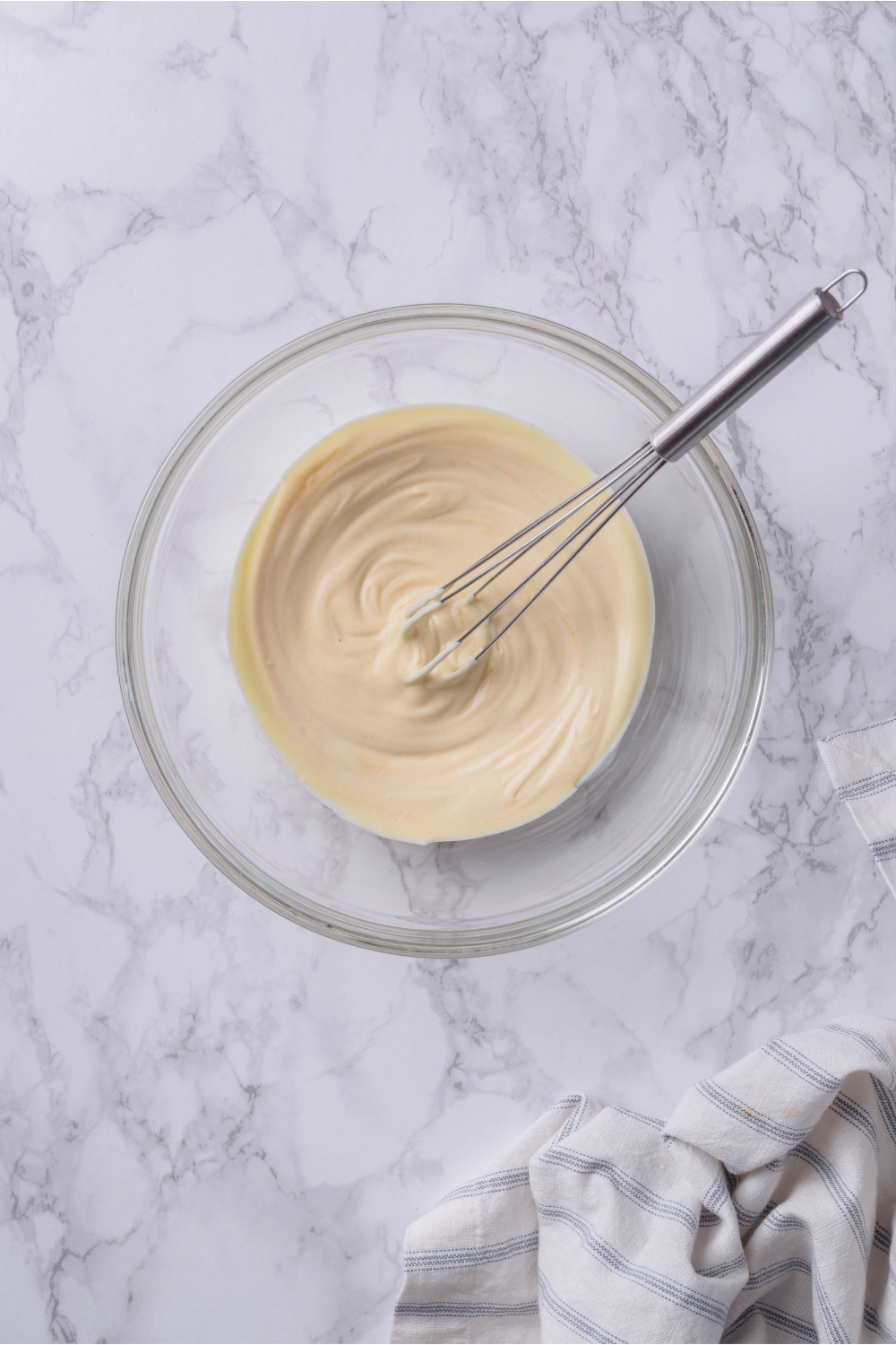A clear bowl of a mayonnaise mixture that's been combined using a whisk that's in the bowl.