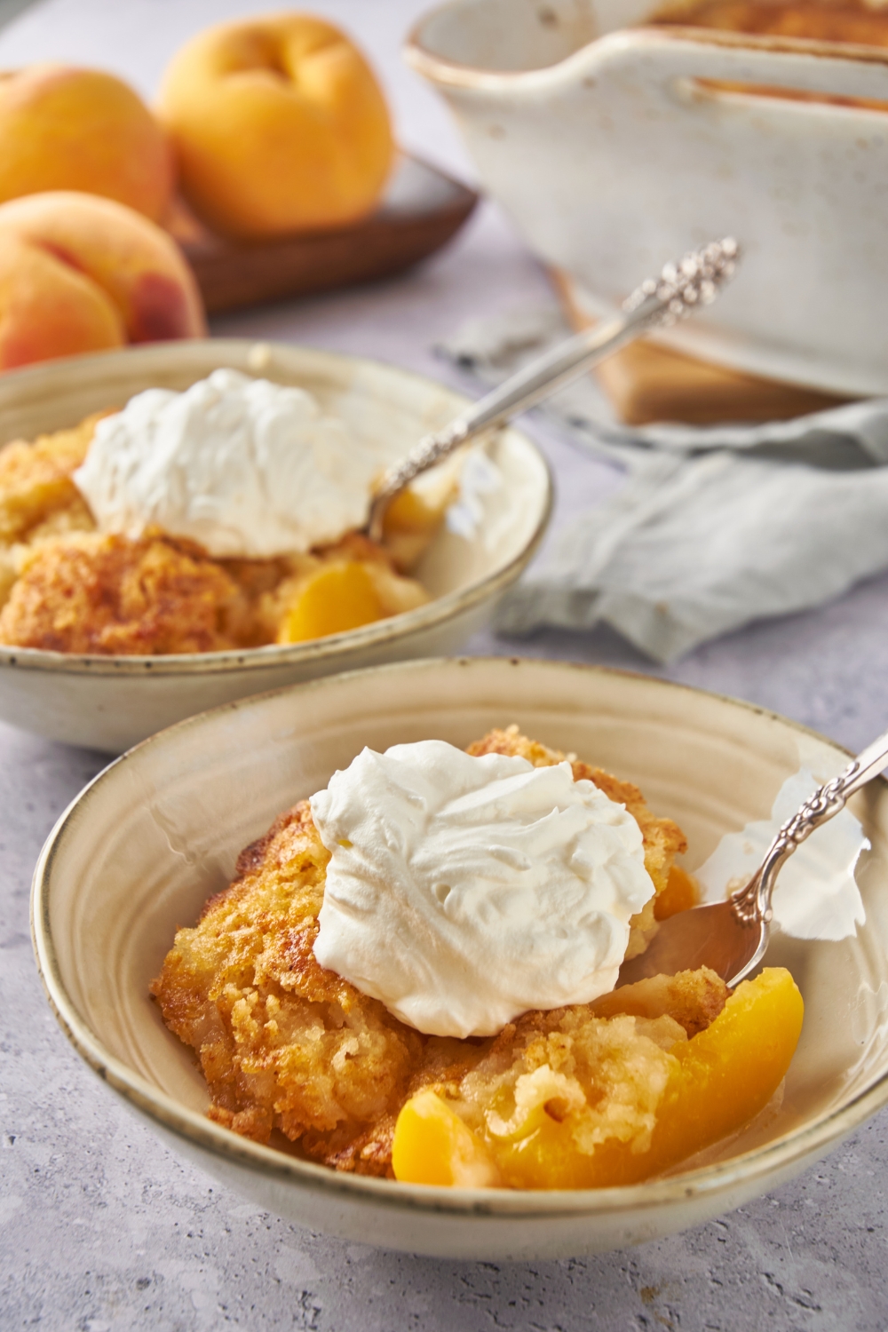 Two bowls of peach cobbler topped with whipped cream and a spoon in each bowl.