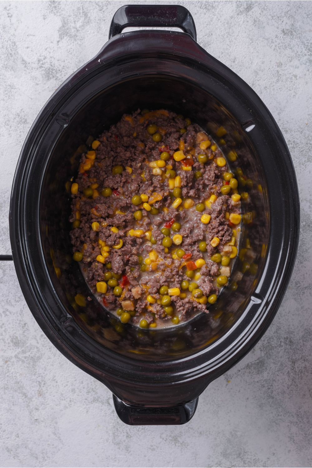 A slow cooker with an even layer of ground beef, corn, peas, and diced peppers.