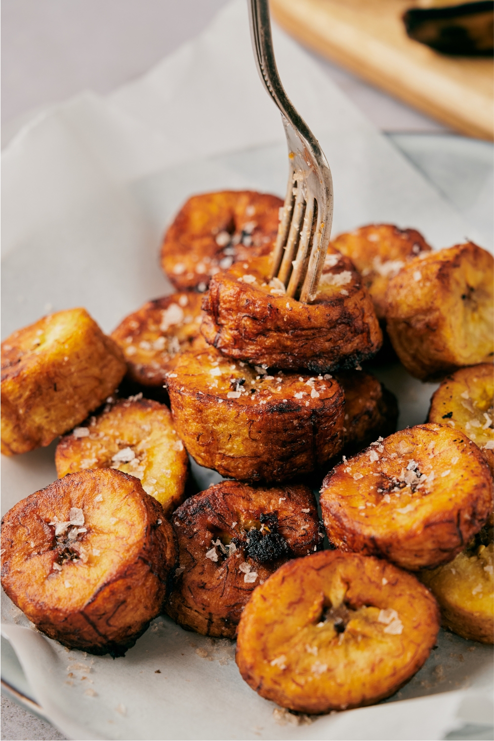 A fork piercing two fried plantain slices off a plate filled with fried plantains, all covered in coarse sea salt.