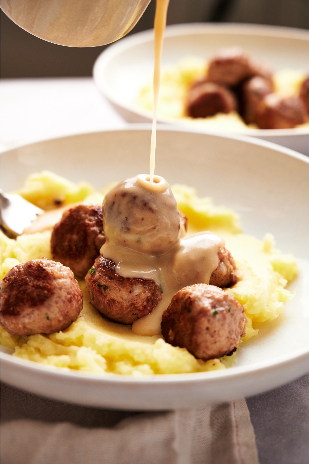 A plate filled with meatballs over top mashed potatoes and a creamy gravy is being poured over top the meatballs.