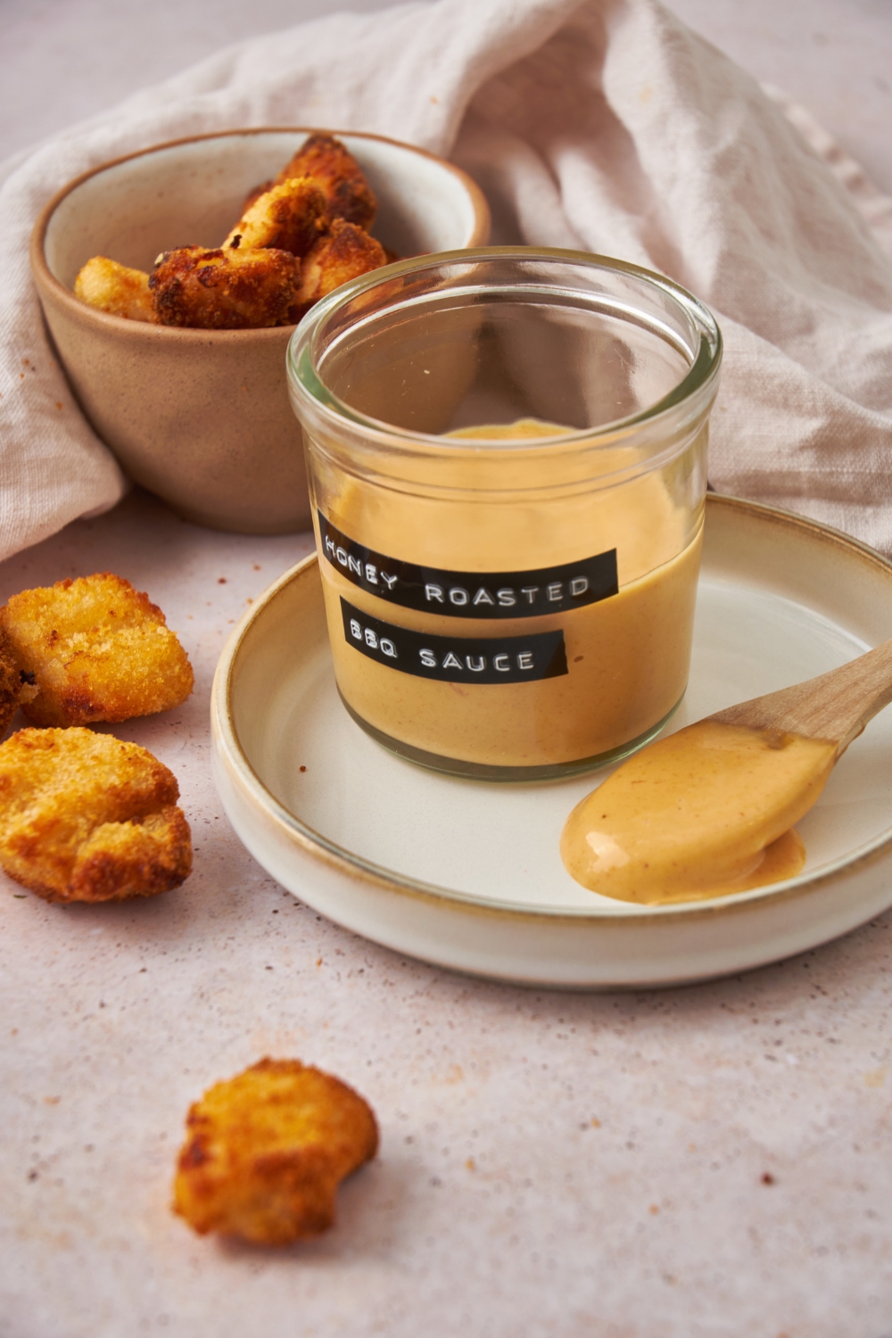 A clear jar labeled "honey roasted BBQ sauce" on a white plate with a spoonful of sauce next to it and three nuggets have fallen on the counter next to the plate.