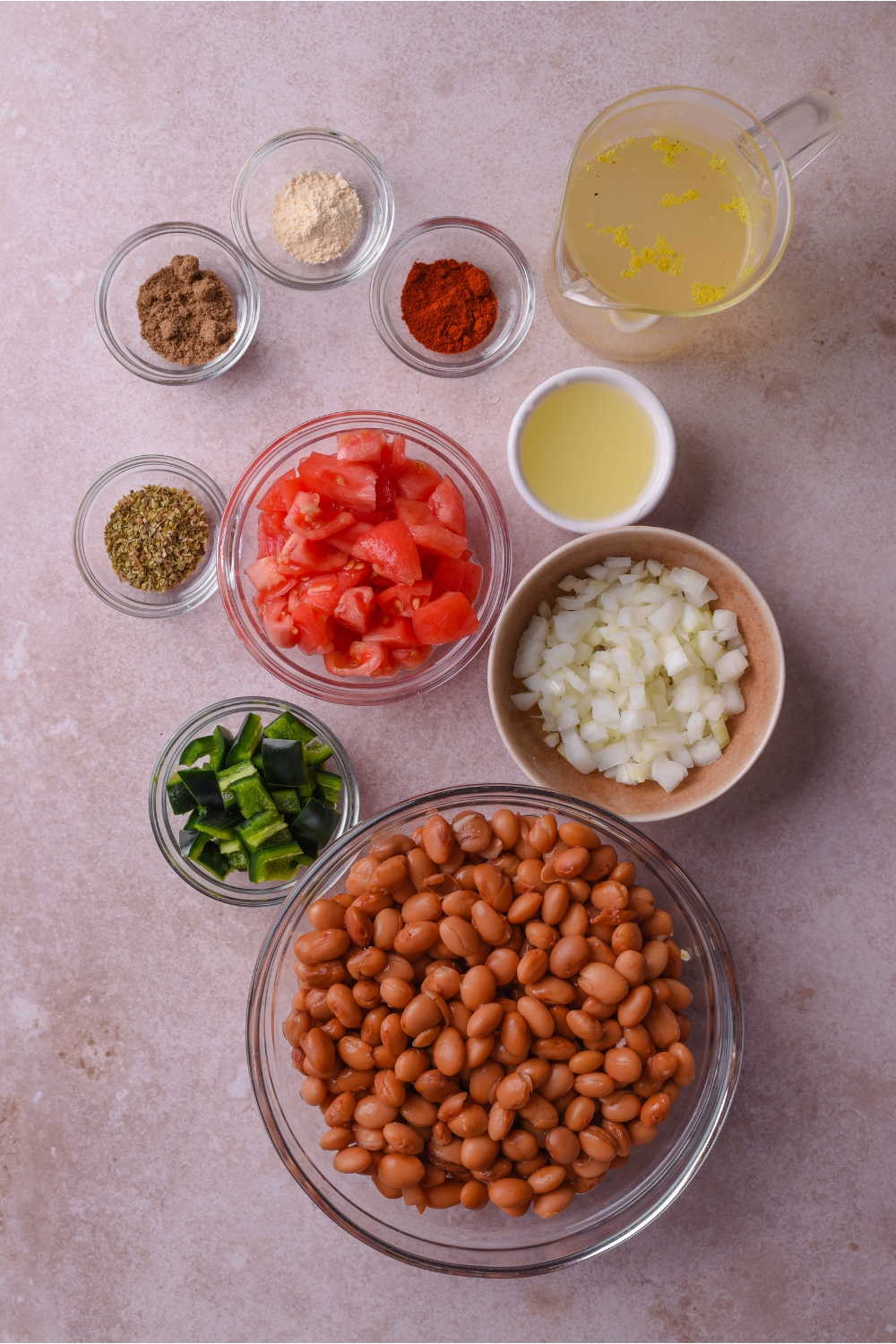 An assortment of ingredients including bowls of pinto beans, broth, diced tomatoes, diced onions, peppers, and spices.