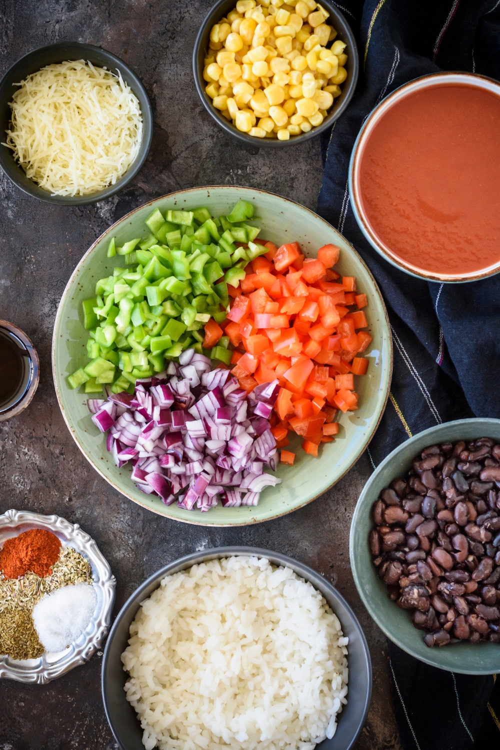 An assortment of ingredients including a bowl of diced peppers and red onion next to bowls of corn, red sauce, white rice, black beans, and shredded cheese.