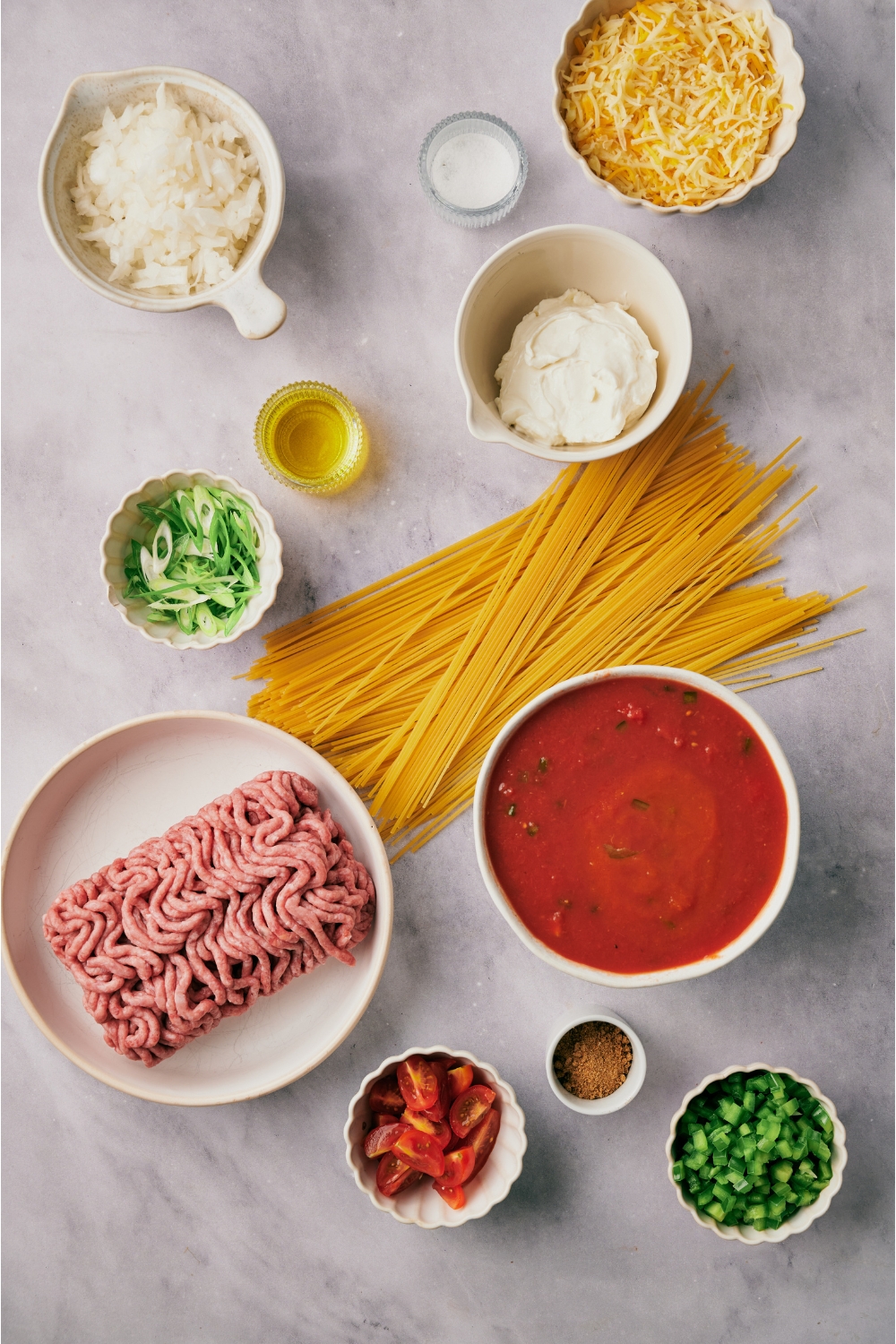 An assortment of ingredients including a pile of dried spaghetti noodles and bowls of tomato sauce, raw ground beef, diced onion, peppers, oil, shredded cheese, and green onion.