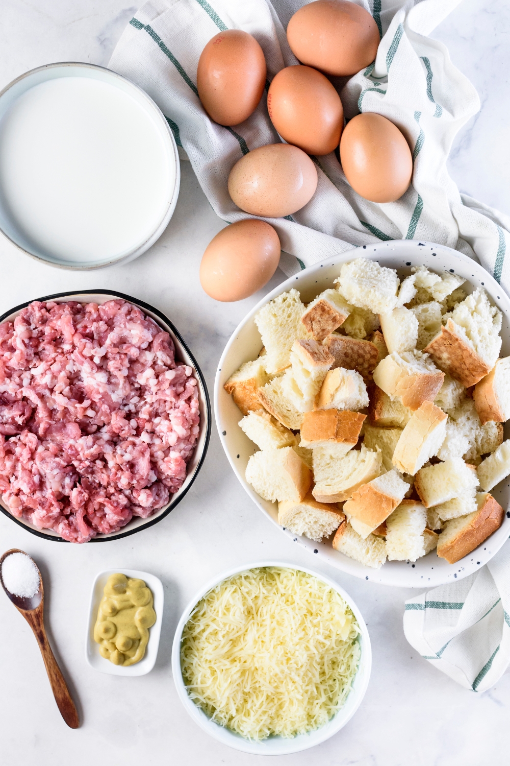 An assortment of ingredients including bowls of raw pork sausage, shredded cheese, mustard, milk, bread cubes, and six eggs wrapped in a dish towel.