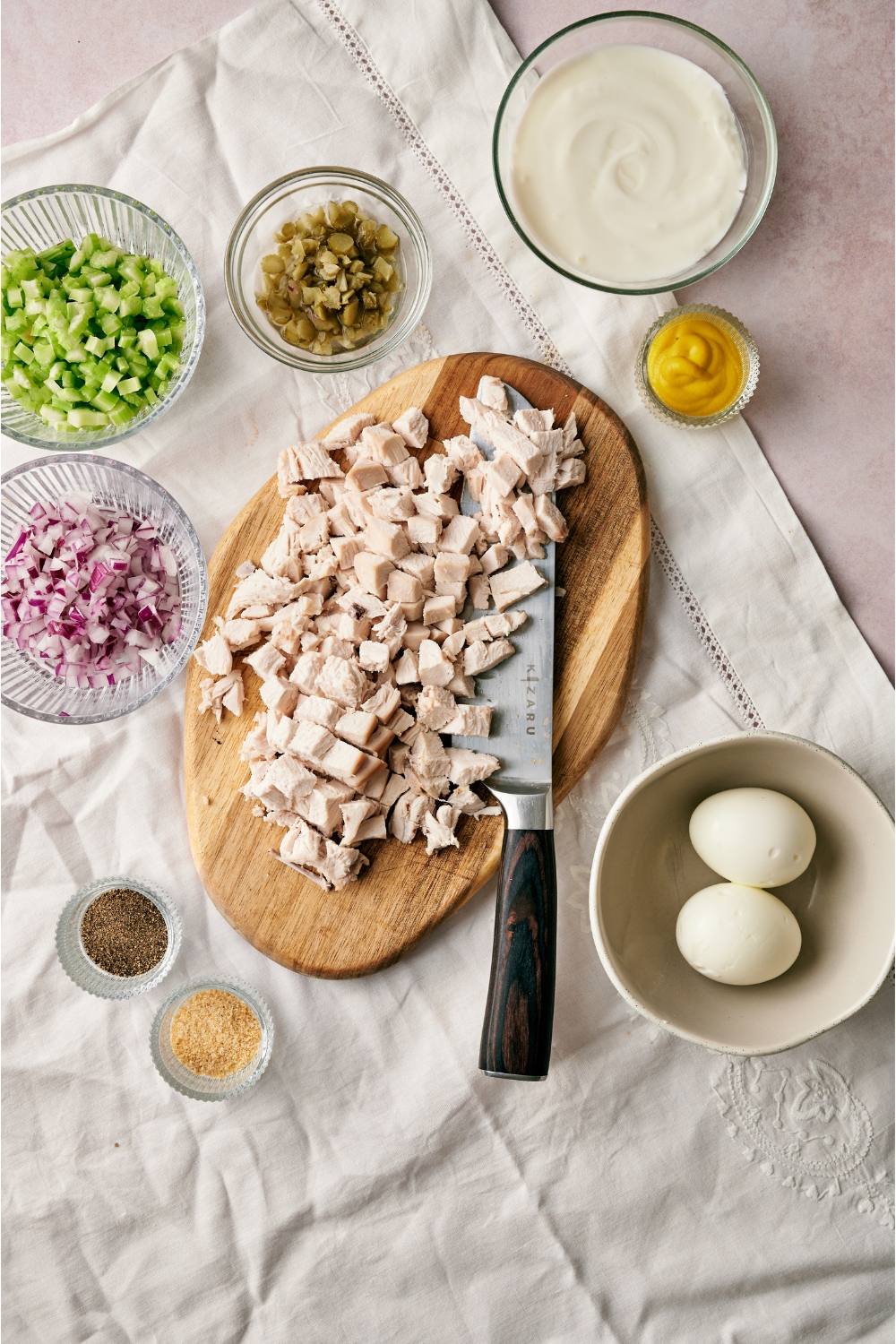 An assortment of ingredients including chopped chicken on a wooden board and small bowls of mayonnaise, mustard, hard boiled eggs, spices, chopped celery, red onion, and pickles.