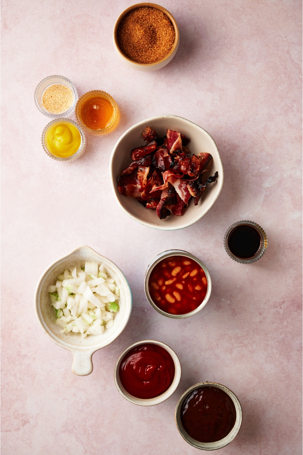 An assortment of ingredients including bowls of diced onions, brown sugar, chopped bacon, ketchup, mustard, barbecue sauce, spices, and a can of beans.