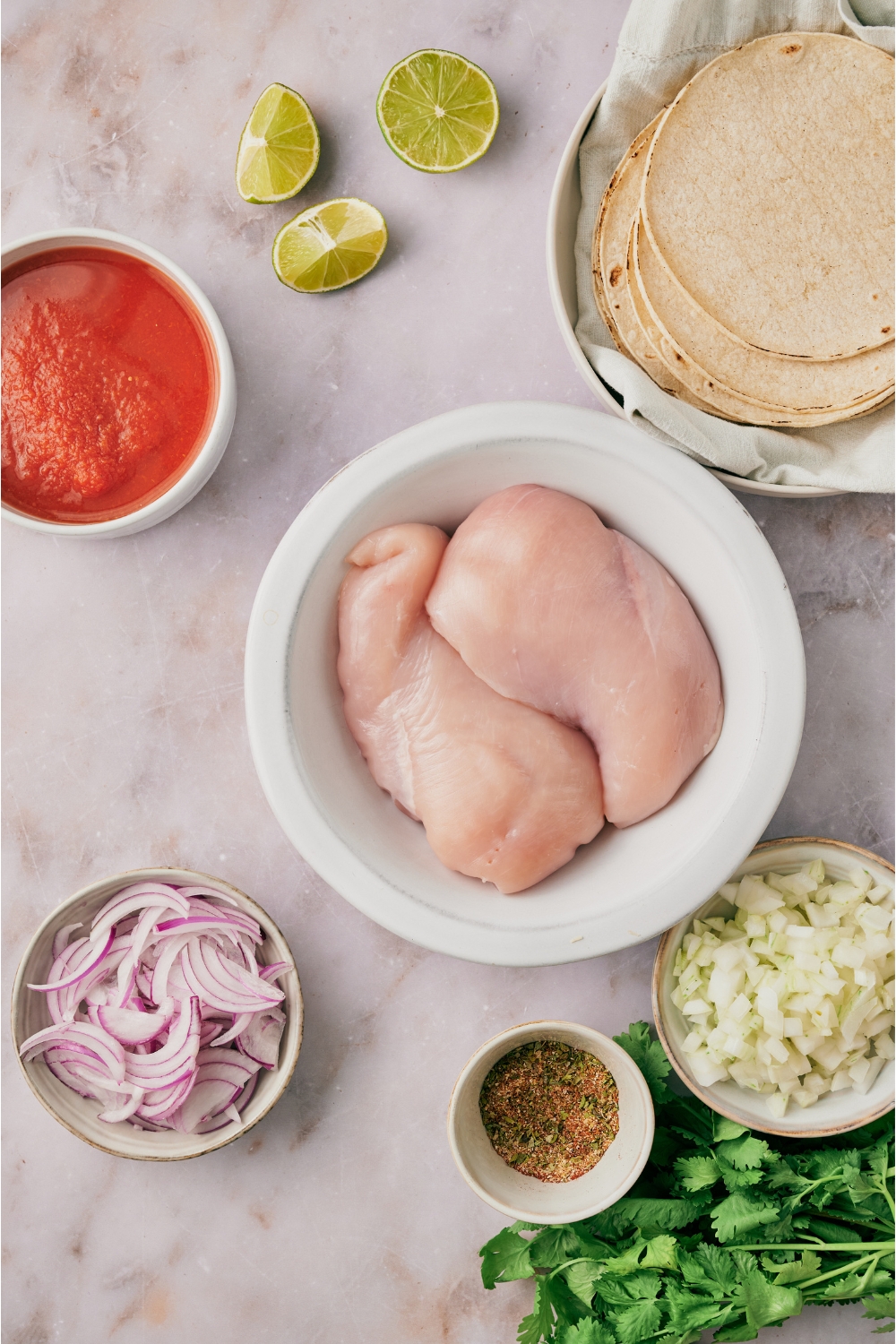 An assortment of ingredients including bowls of raw chicken breasts, sliced red onion, diced white onion, tomato sauce, spices, a plate of tortillas, and lime wedges.