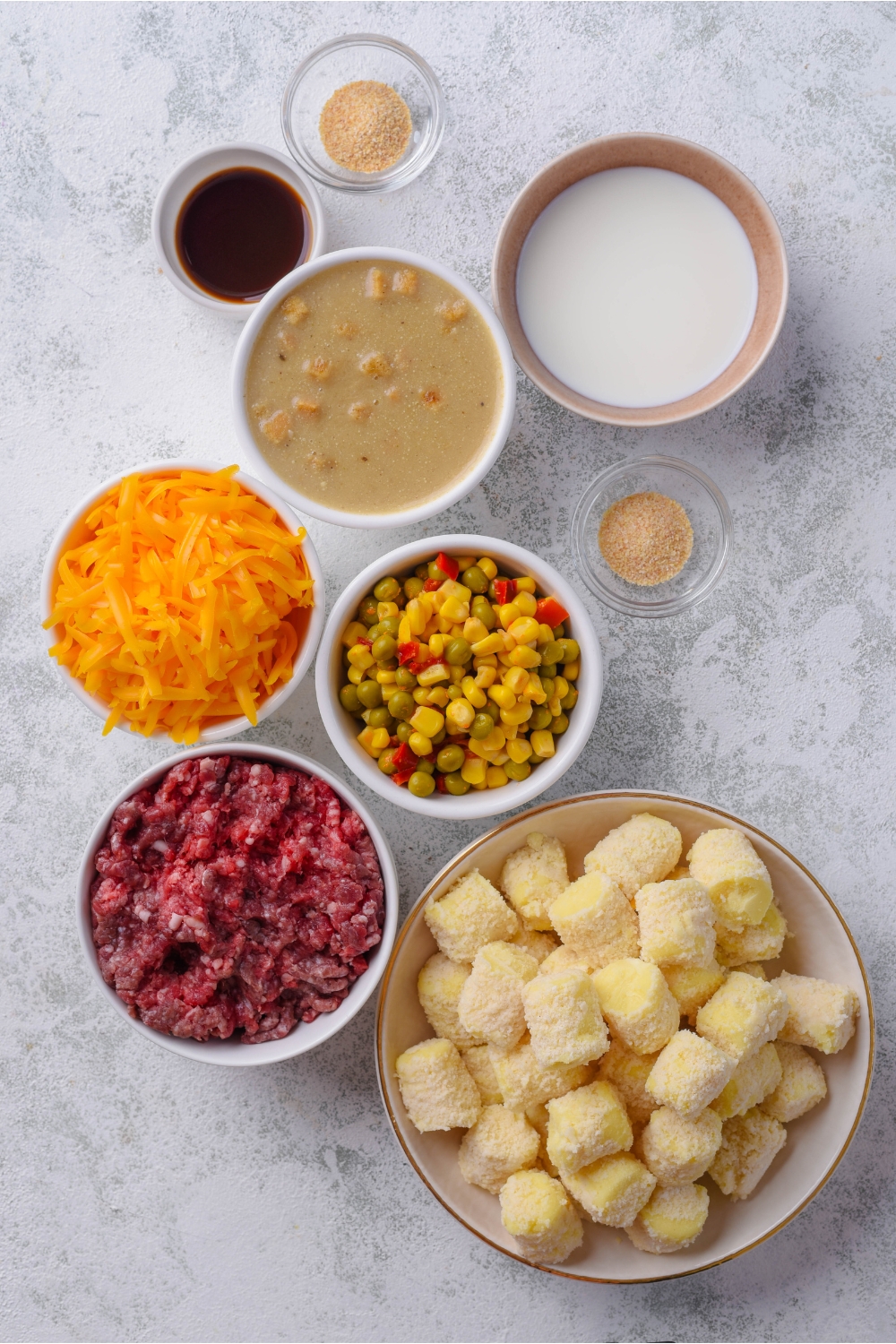 An assortment of ingredients including bowls of frozen tater tots, raw ground beef, shredded cheese, milk, creamy soup, mixed vegetables, and spices.