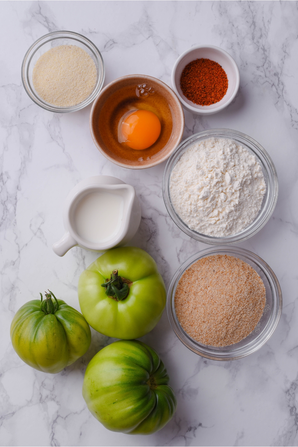 An assortment of ingredients including three green tomatoes, a pitcher of milk, and bowls of flour, bread crumbs, paprika, and an unbeaten egg.