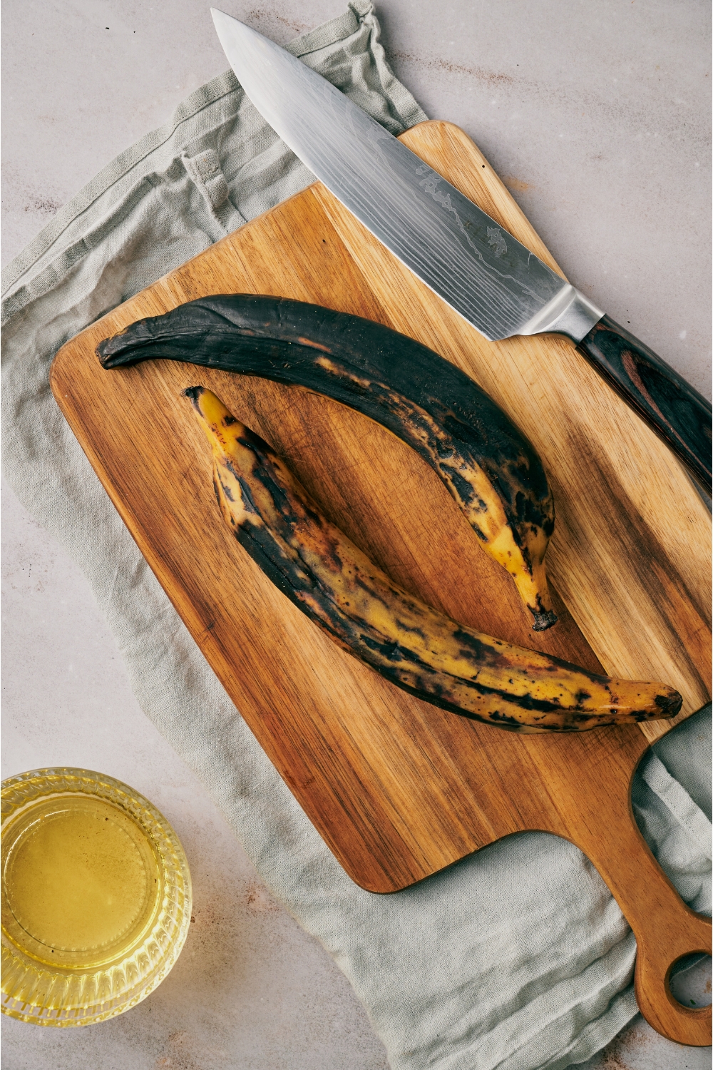 A wood cutting board with a sharp knife and two very ripe plantains on it. The board is next to a small dish of oil.