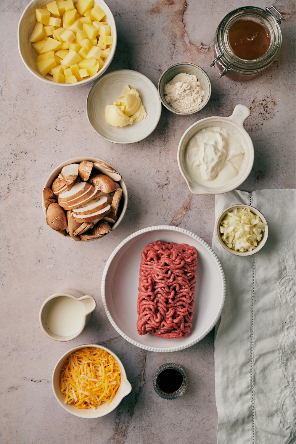 An assortment of ingredients including bowls of diced potatoes, raw ground beef, sliced mushrooms, sour cream, butter, cheese, diced onion, and a jar of beef broth.
