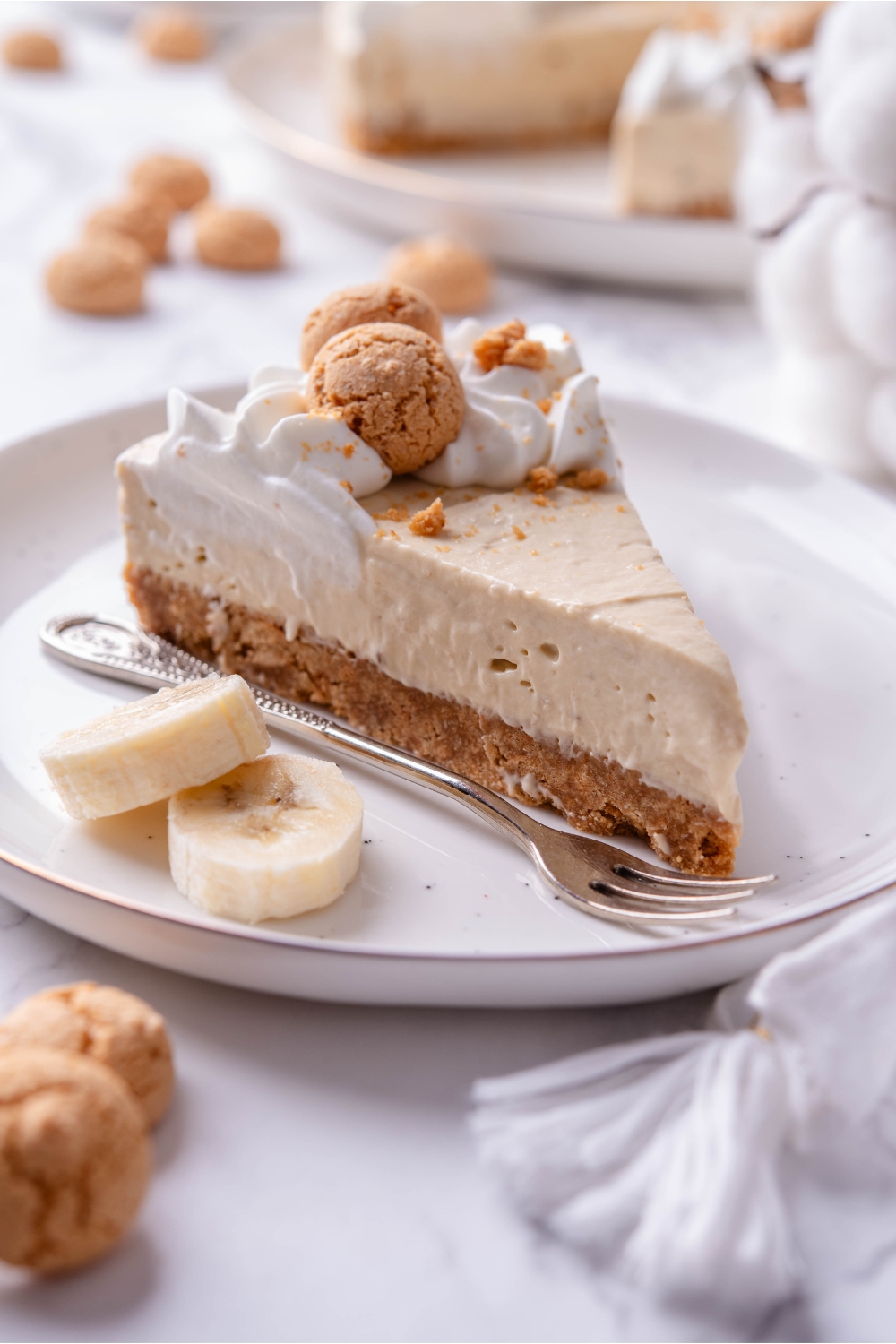 A serving of banana cheesecake on a plate with two banana slices and a fork. The cheesecake has a cookie crust and is topped with whipped cream and mini cookies.