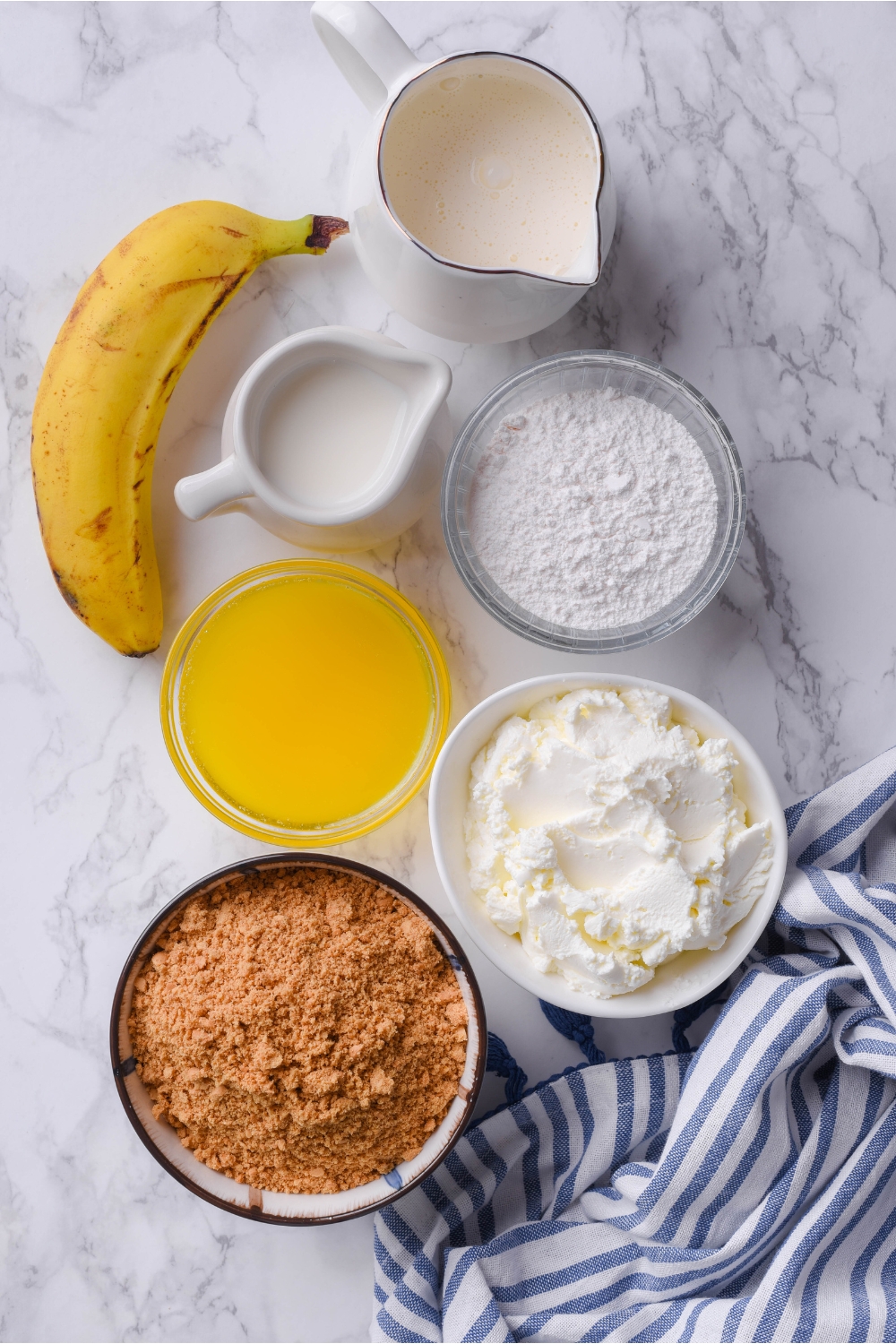 An assortment of ingredients including bowls of cream cheese, pudding mix, melted butter, cookie crumbs, jars of cream and milk, and a single banana.