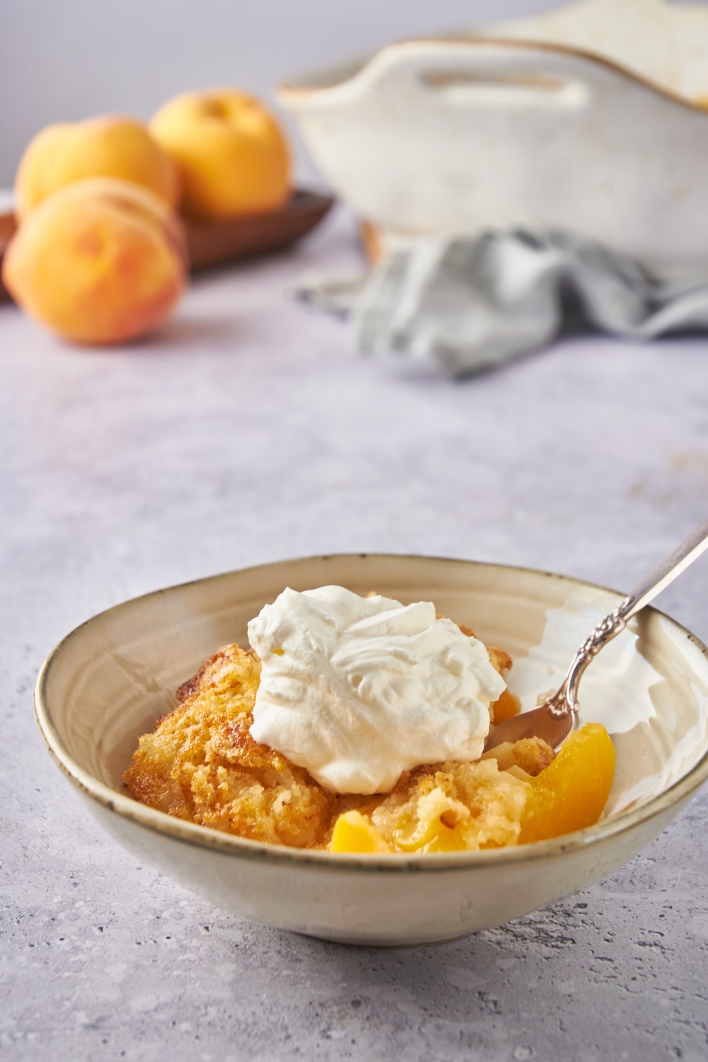 A bowl of peach cobbler topped with whipped cream and a spoon is in the bowl.