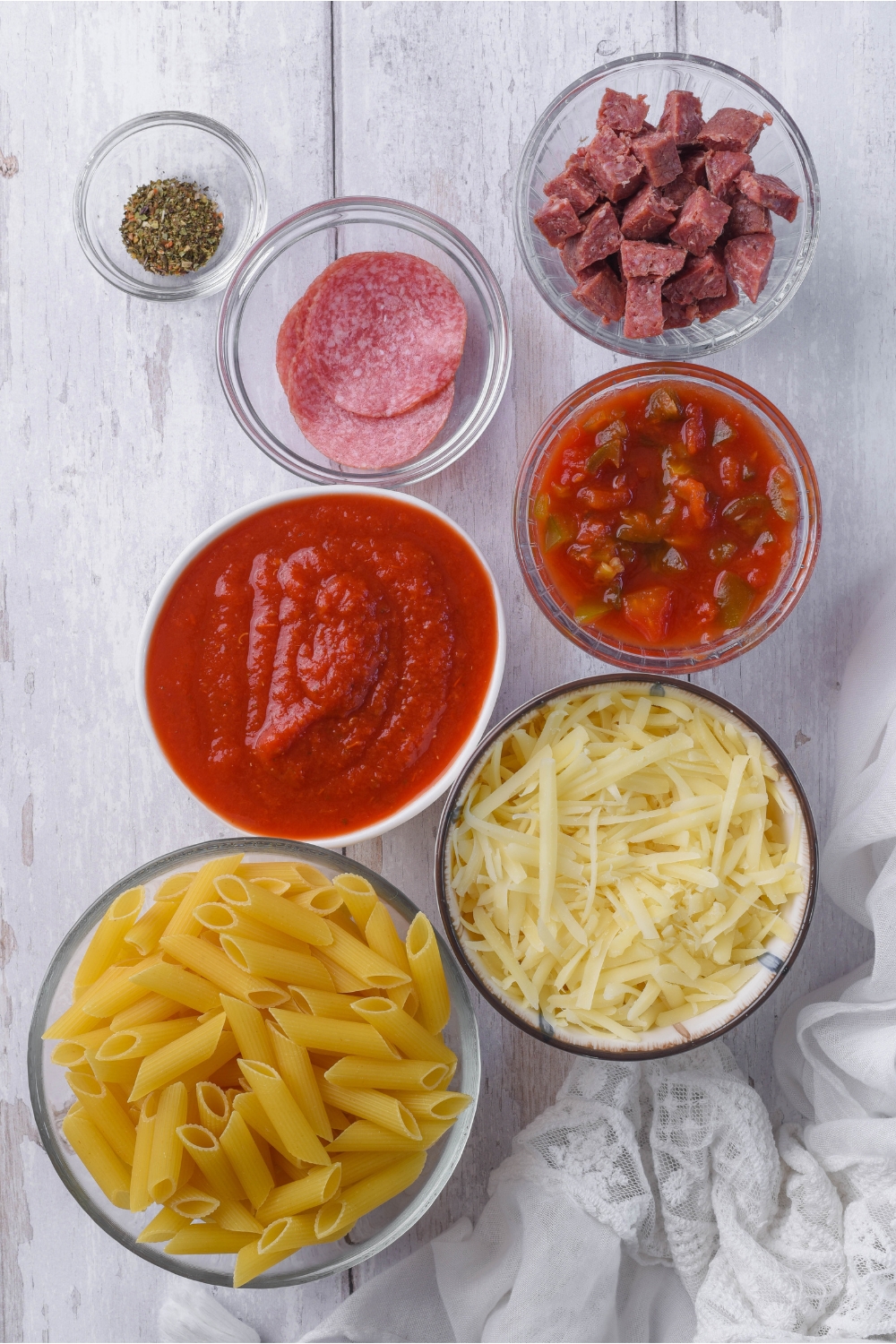 An assortment of ingredients including bowls of red sauce, diced pepperoni, sliced pepperoni, shredded cheese, spices, and dried pasta.