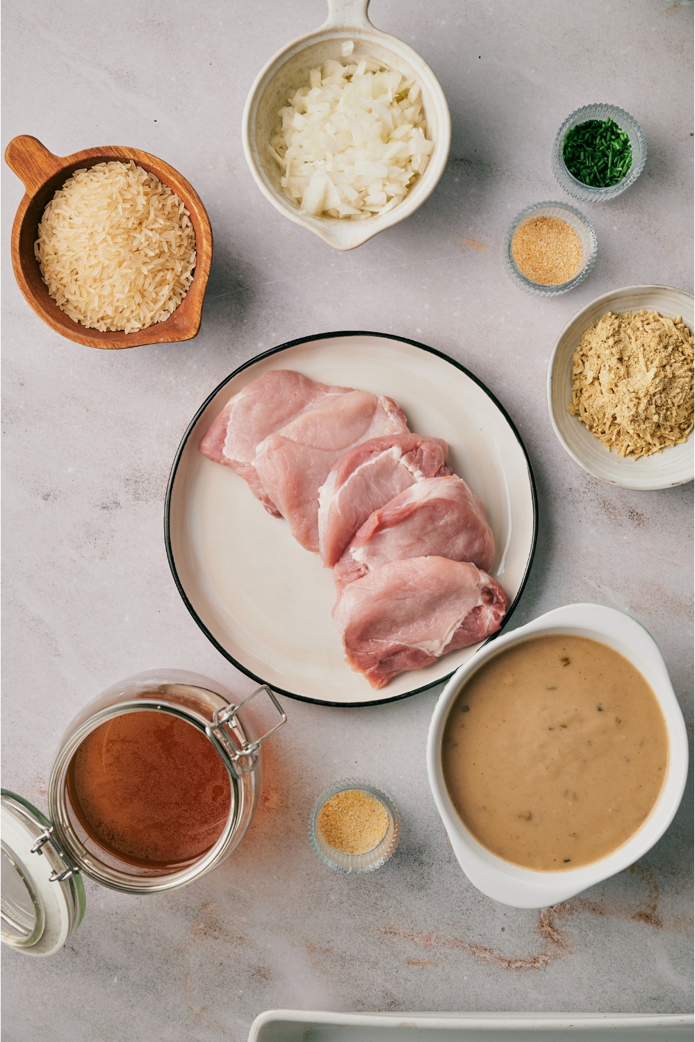 An assortment of ingredients including a plate of pork chops and bowls of mushroom soup, shredded cheese, diced onion, broth, and spices.