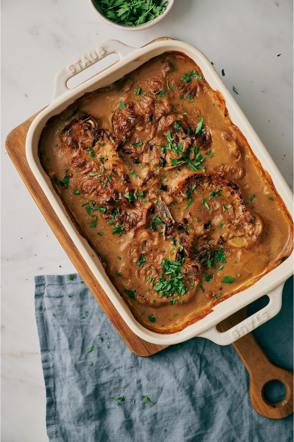 A white baking dish filled with browned pork chops smothered in a creamy sauce and garnished with fresh parsley.