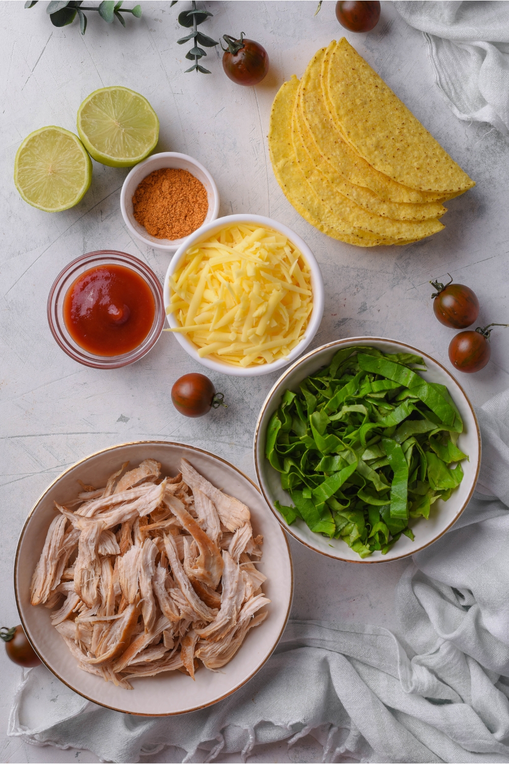 An assortment of ingredients including a stack of crispy taco shells, a lime cut in half, cherry tomatoes, and bowls of tomato paste, shredded lettuce, shredded cheese, spices, and shredded chicken.