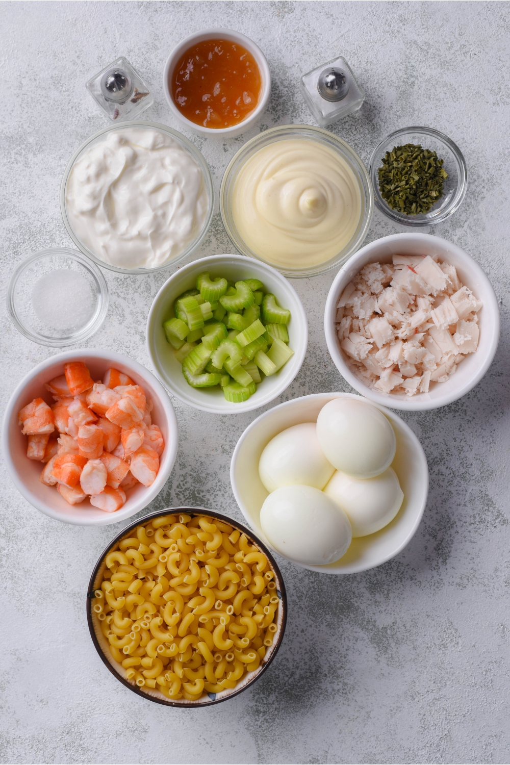 An assortment of ingredients including bowls of dried pasta, hard-boiled eggs, shrimp pieces, shredded crab meat, mayonnaise, diced celery, sour cream, herbs, and spices.