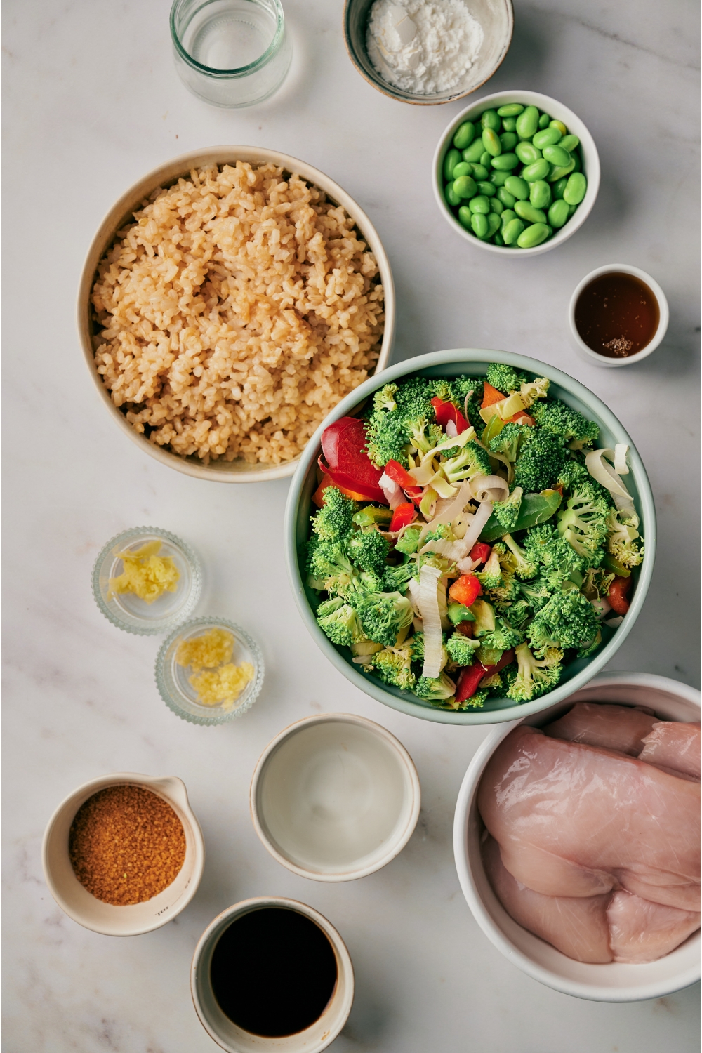An assortment of ingredients including bowls of raw vegetables, cooked brown rice, edamame, minced garlic, minced ginger, brown sugar, soy sauce, and raw chicken breasts.