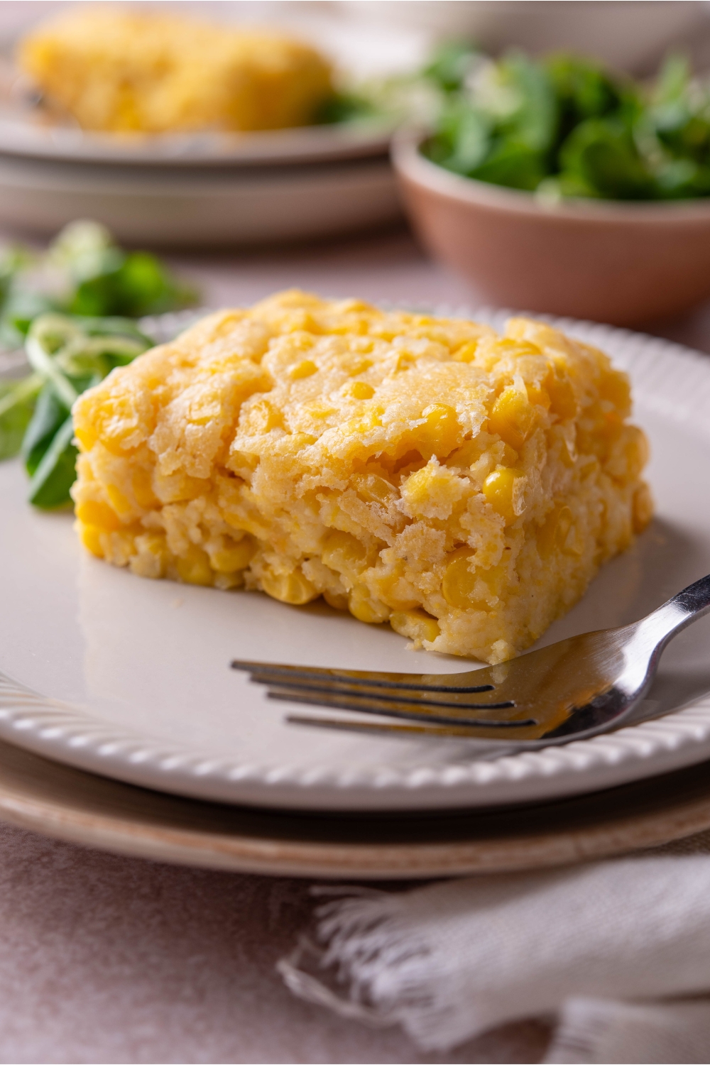 A square of corn casserole with a fork and a pile of fresh greens on the plate.