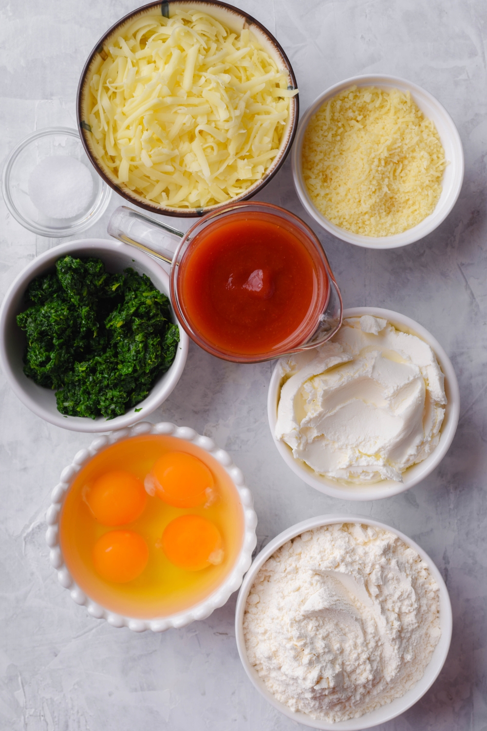 A countertop with all the ingredients to make manicotti crepes. Parmesan cheese, mozzarella cheese, ricotta cheese, salt, marinara sauce, thawed and drained chopped spinach, cracked eggs, and flour are all in separate bowls on the countertop.