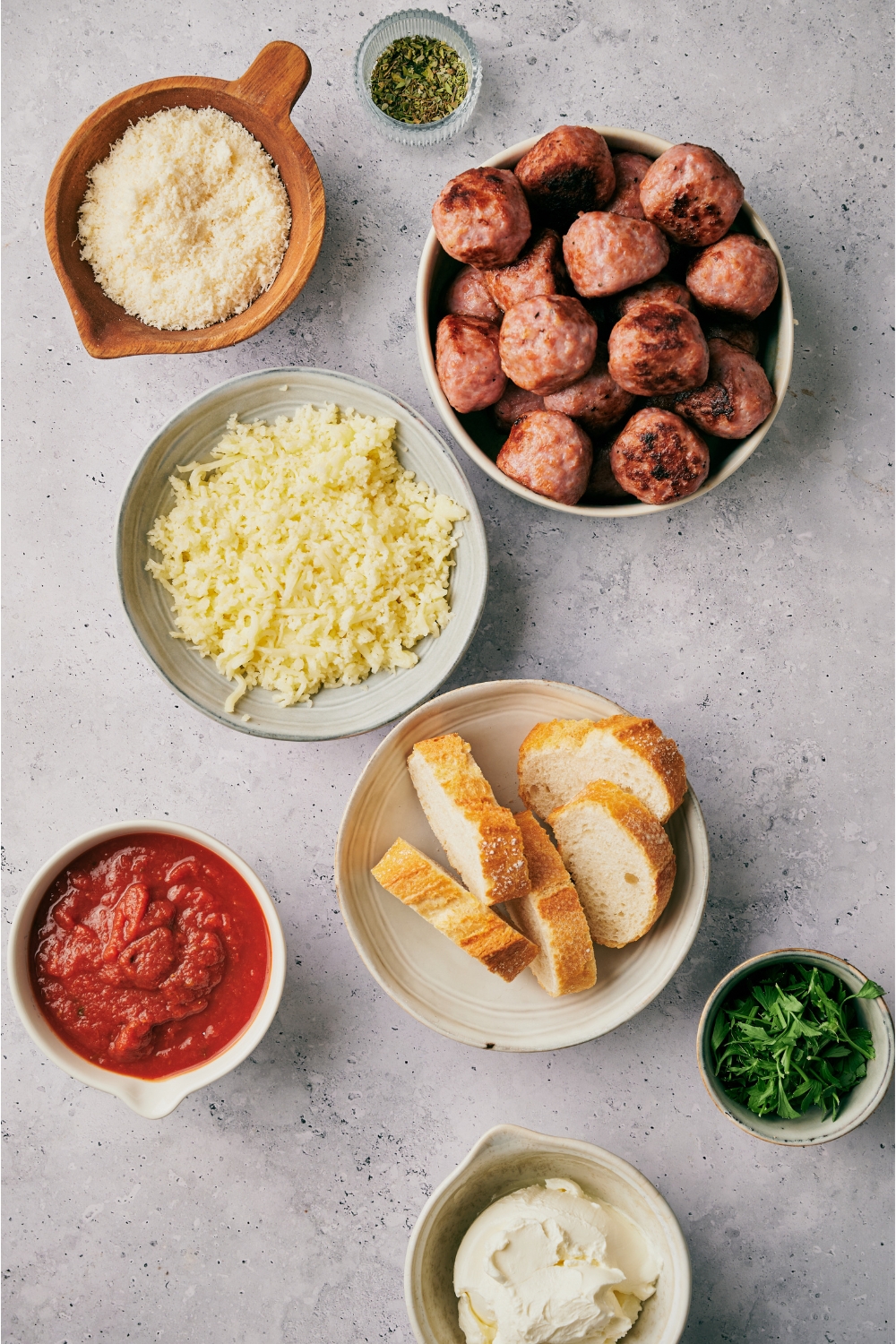An assortment of ingredients including bowls of shredded cheese, bread slices, meatballs, red sauce, cream cheese, and herbs.