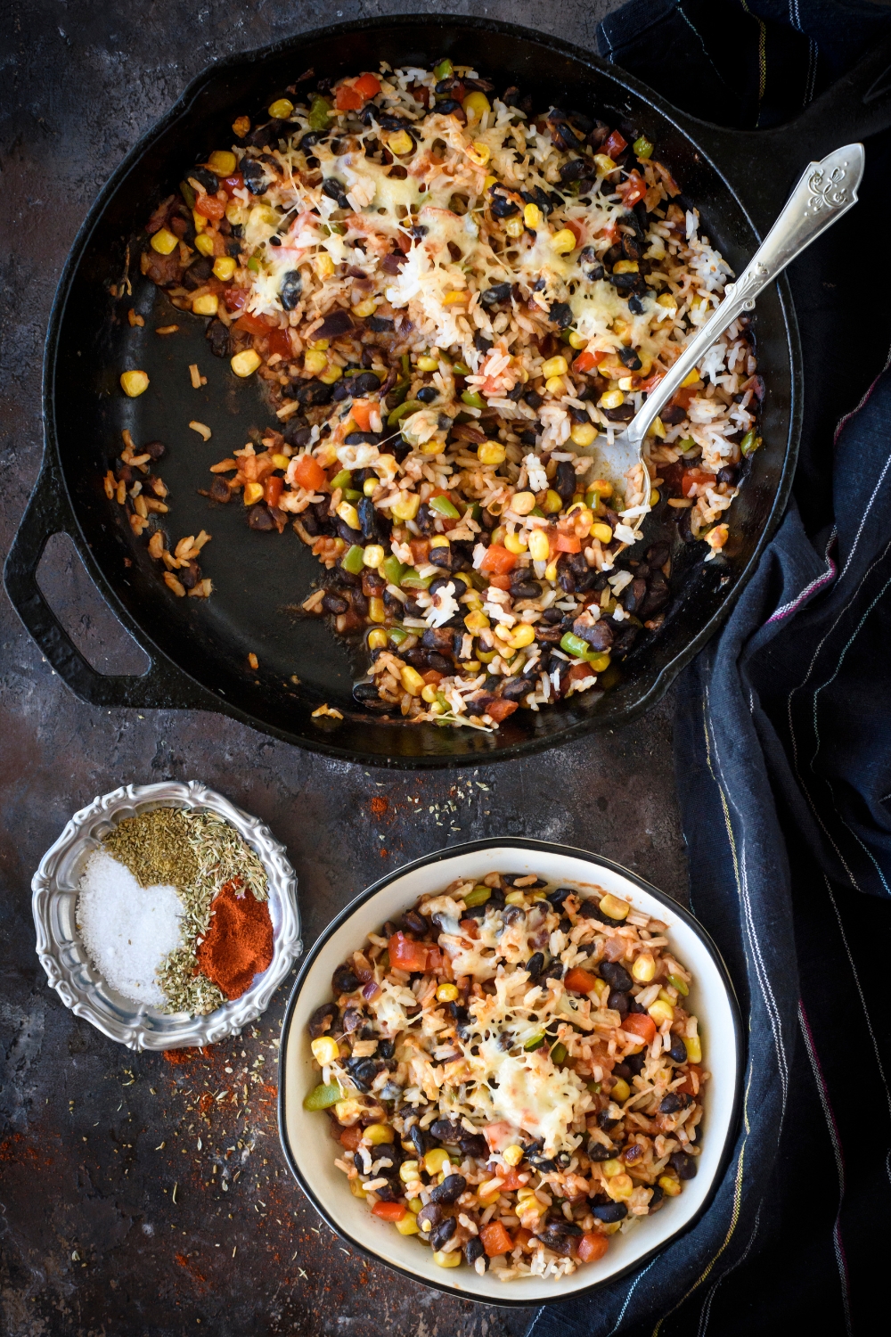 A skillet half filled with rice casserole, with one serving scooped out and served in a bowl next to the skillet. There is also a bowl of spices on the table.