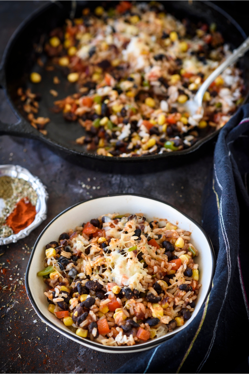 A bowl of rice casserole covered in melted cheese with visible chunks of vegetable, corn, and black beans. The rest of the rice casserole is in the background in a skillet.