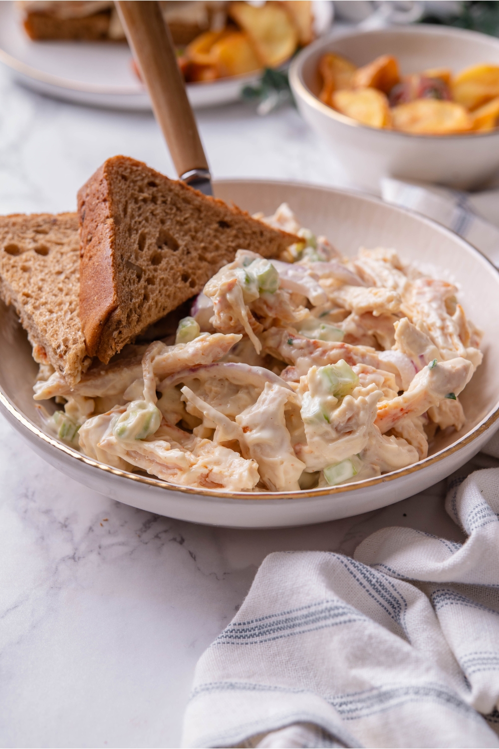 A bowl of chicken salad with a piece of whole wheat bread sliced in half in the bowl along with a fork.