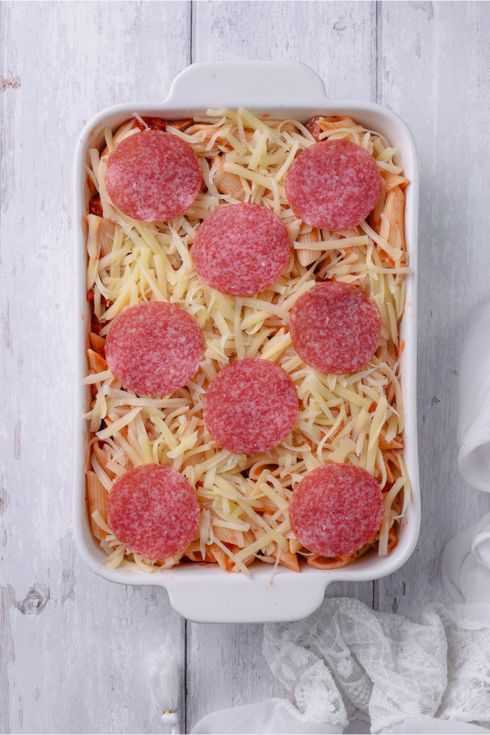A white baking dish filled with unbaked pasta in red sauce, shredded cheese, and a layer of sliced pepperoni on top.