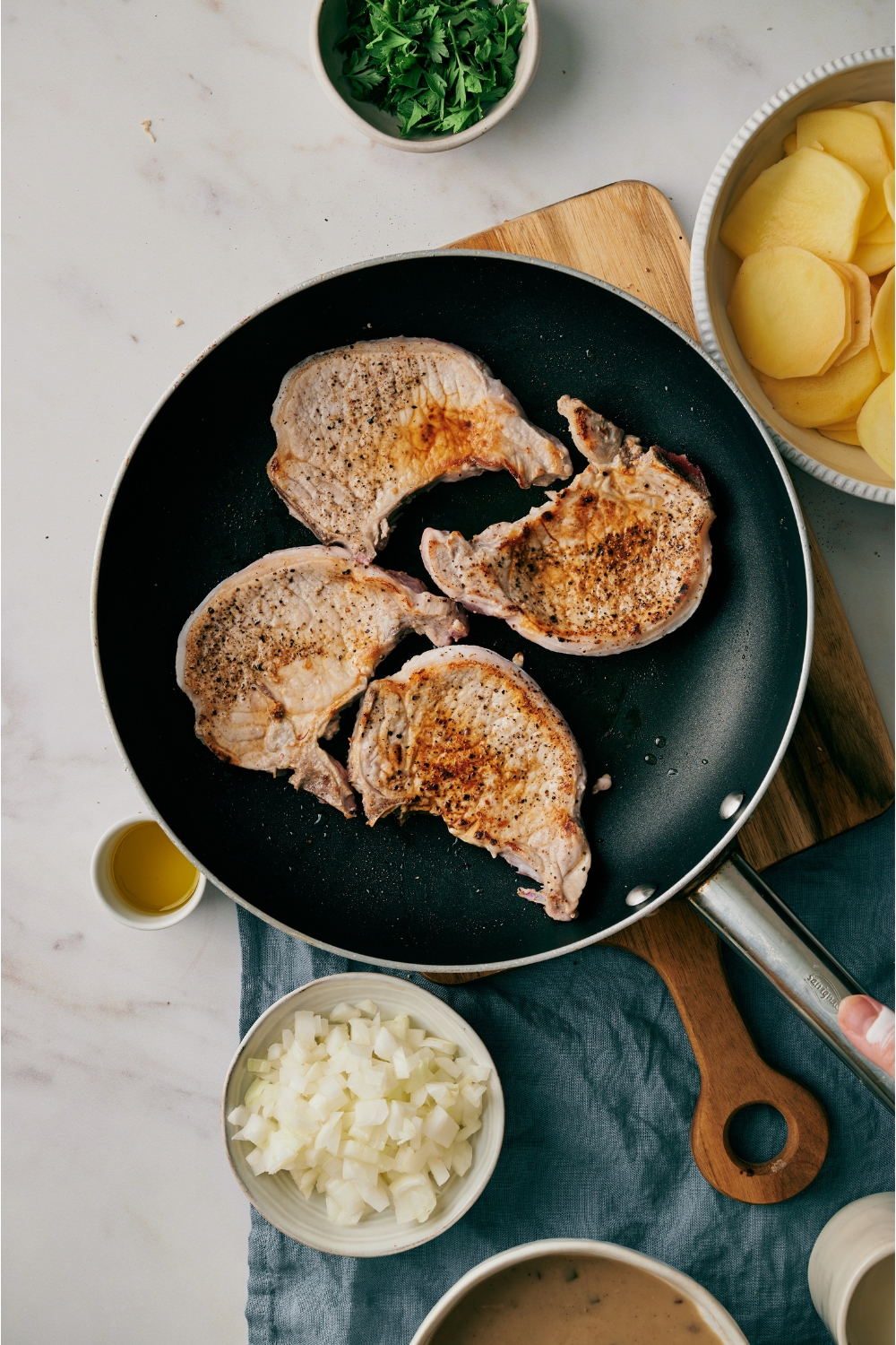 Four boneless pork chops being seared on a black skillet that's surrounded by an assortment of ingredients.