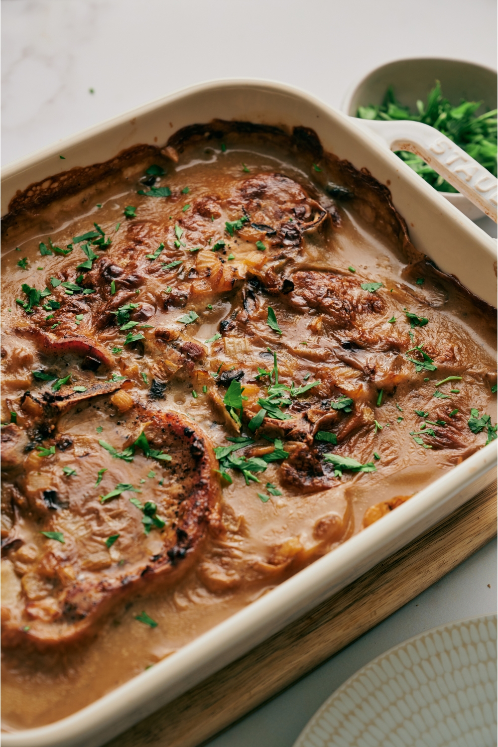 A white baking dish filled with browned pork chops smothered in a creamy sauce and garnished with fresh parsley.