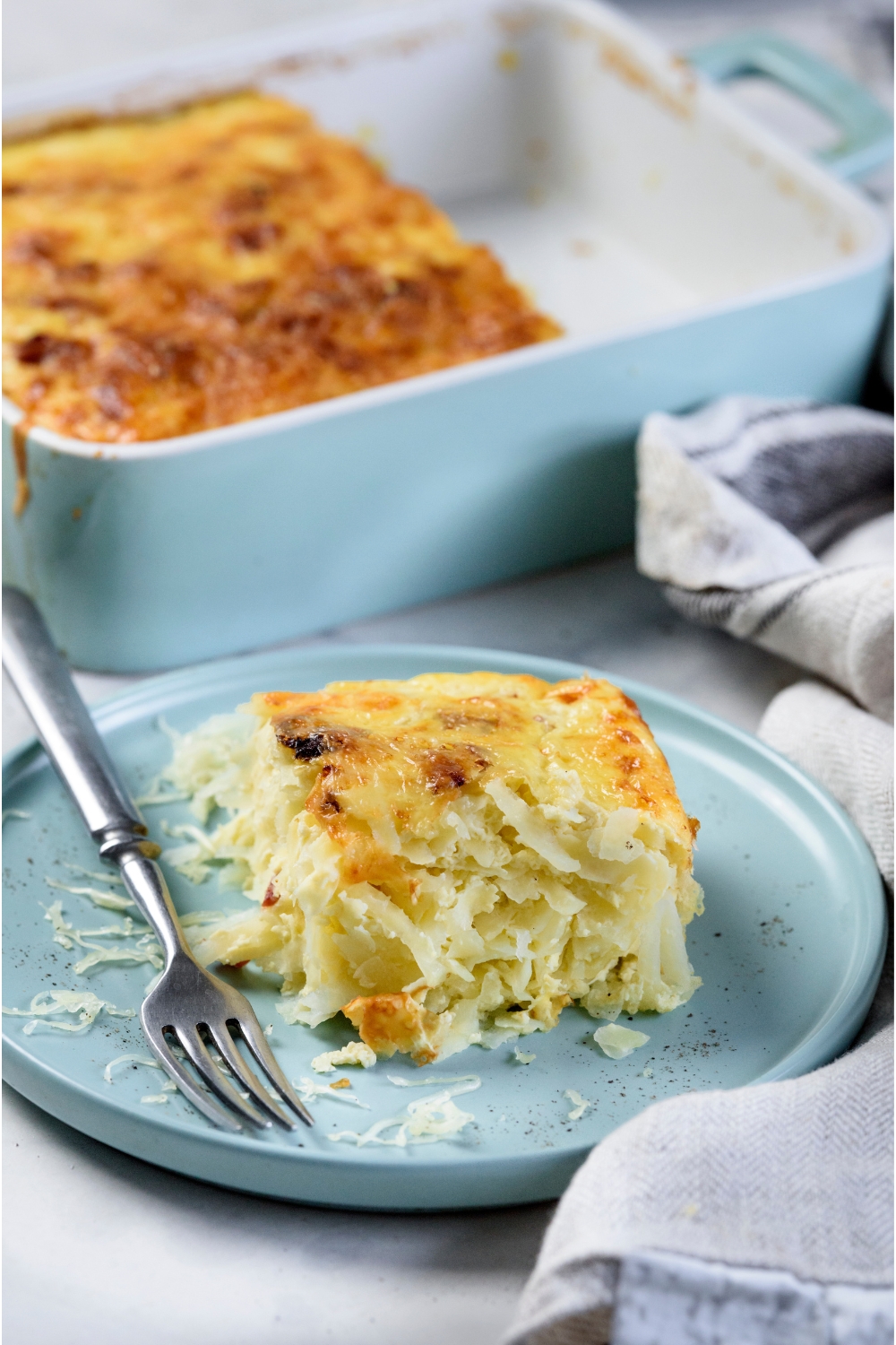 A square serving of Amish breakfast casserole on a blue plate with a fork on the plate and the rest of the casserole in the background.